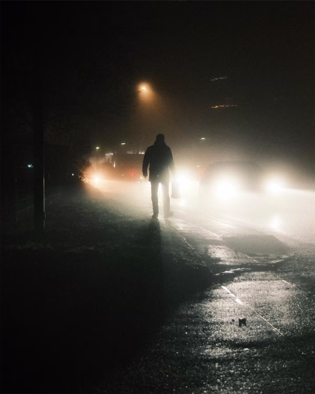 A man walks along a wet sidewalk at night, lit from behind by car headlights and a street light in the fog, as shot